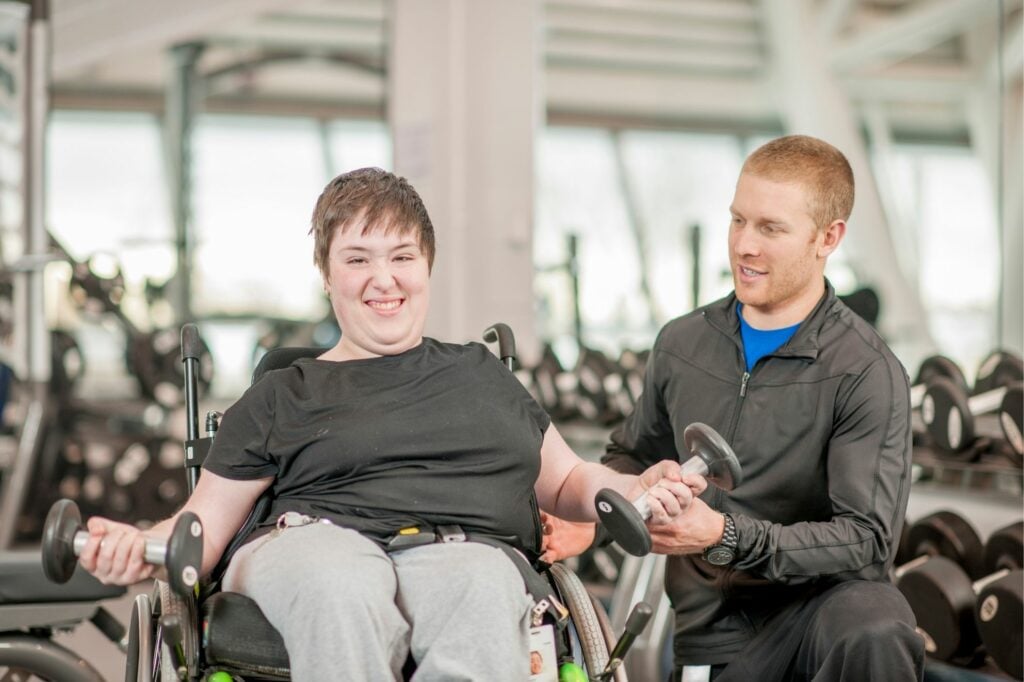 Smiling wheelchair user using dumb bells with a fitness coach